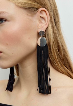 Donna bionda che indossa grandi orecchini circolari d'argento con lunghe nappine nere che pendono fino alla spalla.