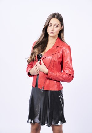 Giacca da motociclista in pelle rossa con zip asimmetrica, indossata su un vestito nero in satin con orlo a campana, su uno sfondo bianco.