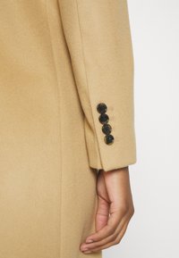 Manica di cappotto in lana beige con bottoni neri e dettagli di cucitura visibili. Il tessuto ha una consistenza morbida e una leggera lucentezza.