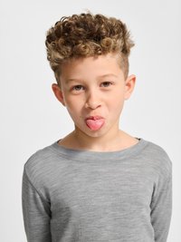 Jongen met krullend haar die een grijs shirt met lange mouwen draagt en zijn tong uitsteekt tegen een effen lichte achtergrond.