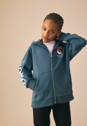 Młoda osoba z warkoczami typu cornrow, ubrana w niebieską bluzę z kapturem i zamkiem marki Pokémon z logo i napisem, pozująca z jedną ręką za szyją.