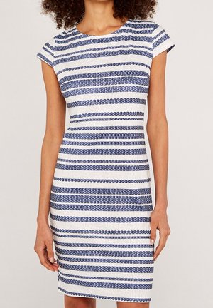 Frau trägt ein figurbetontes weißes Kleid mit abwechselnden horizontalen dunkelblauen Streifen, die aus kleinen kreisförmigen Mustern bestehen.