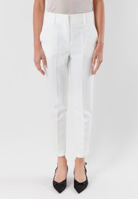 Pantaloni bianchi su misura con pieghe, vestibilità slim e tasche laterali. Il tessuto ha una consistenza liscia. Abbinati a décolleté neri a punta.