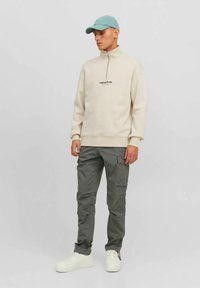 Beige neljännesvetoketjullinen collegepaita, jossa pieni musta logo, yhdistetty vihreisiin cargo-housuihin ja valkoisiin tennareihin. Asusteina vaaleansininen lippalakki.