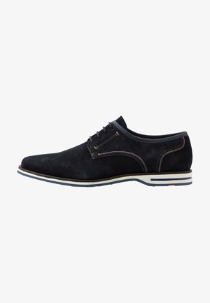 Chaussure en daim noir à finition lisse, coutures claires contrastantes et semelle en caoutchouc avec une subtile bande blanche et bleue.