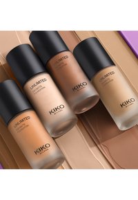 Quatre flacons de fond de teint mat KIKO Milano Unlimited 24H avec des bouchons noirs, présentant différentes teintes de beige sur un fond texturé.