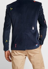 Granatowa aksamitna marynarka z świątecznymi haftami, w tym ostrokrzew, bałwanek, laski cukrowe i prezent. Klasyczny krój z akcentami guzików.