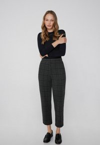 Zwarte langemouwen top gecombineerd met grijs geruite broek met rode en groene lijnen, cropped pasvorm en zwarte loafers op blote voeten.
