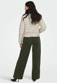 Sieviete ar gariem tumšiem matiem, tērpusies gaiši bēšā pufīgajā jakā, olīvzaļās platās biksēs un melnās augstpapēžu zābakos, stāv ar muguru vērsta.