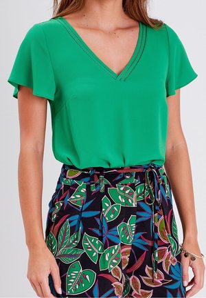 Vrouw draagt een groene korte mouw V-hals top, ingestopt in een broek met hoge taille met een kleurrijke tropische bladerenprint en een gestrikte tailleriem.