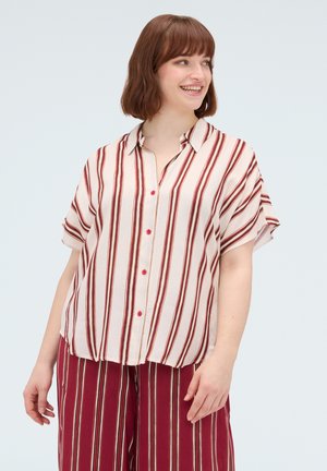 Vrouw met kort bruin haar die glimlacht, draagt een loszittend crème shirt met verticale rode strepen en bijpassende rood gestreepte broek.