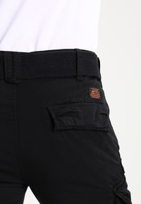 Zwarte cargobroek met een gestructureerde riem, ritszakken en een klein oranje label op de achterzak. Duurzaam materiaal met een functioneel ontwerp.