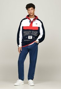 Chaqueta de sudadera en marino, rojo y blanco con cremallera, gran bolsillo frontal y texto de logo. Combinada con vaqueros azules y zapatillas blancas.