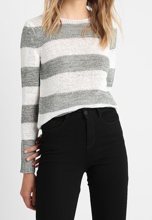 Femme portant un pull tricoté rayé gris et blanc à manches longues et un pantalon noir taille haute, debout devant un fond uni.