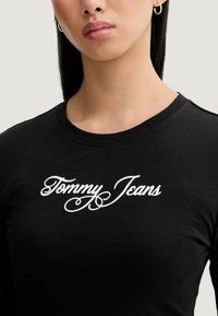Czarna bawełniana koszulka z długim rękawem, z białym logo "Tommy Jeans" w stylu pisanym na wysokości klatki piersiowej. Gładka faktura i dopasowany krój.