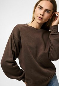 Sudadera marrón con un corte relajado y escote ancho, que presenta mangas largas y una tela suave y texturizada. Con detalles de costura sutiles.