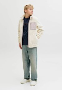 Αγόρι με κοντά ξανθά μαλλιά στέκεται κοιτώντας προς τα αριστερά, φορώντας μια κρεμ φλις ζακέτα, σκούρο μπλουζάκι, φαρδιά μπλε τζιν και λευκά αθλητικά παπούτσια με φόντο λευκό.