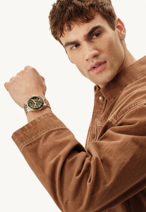 Sølvfarvet metal armbåndsur med en mørkegrøn urskive, båret på en lys brun kordfløjls skjorte med opkædede ærmer. Detaljer inkluderer et rundt urhus og guldmarkører.