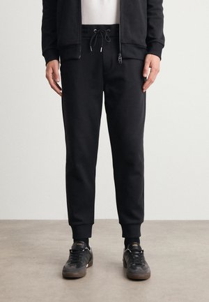 Schwarze Jogginghose aus weichem Stoff mit elastischem Bund, versehen mit Kordeln, seitlichen Taschen und schmal zulaufenden Knöcheln.