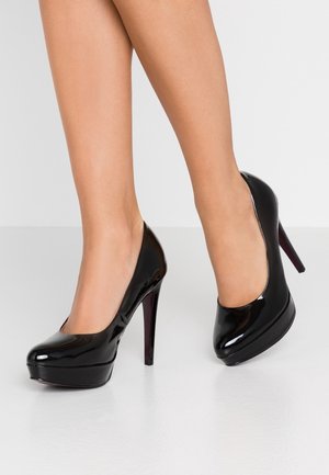 Nogawice ubrane w czarne lakierowane szpilki na platformie stojące na białej podłodze na tle jednolitego tła.