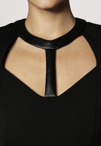 Robe noire présentant un design découpé avec un col en cuir circulaire et une sangle verticale centrale. Tissu lisse avec une texture mate.