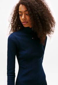 Maglia a collo alto a maniche lunghe color navy con texture a coste; presenta un piccolo logo sul colletto. Design aderente.