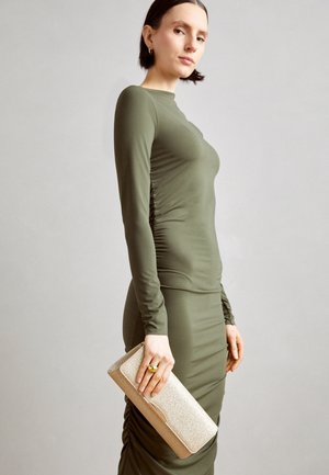 Femme en robe moulante vert olive à manches longues tenant une pochette dorée scintillante sur un fond uni gris clair.