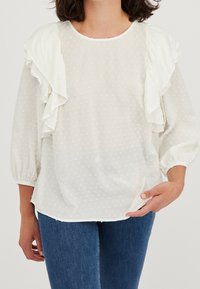 Valkoinen, teksturoidulla pinnalla varustettu blouse, jossa on rypytetyt hihat, pyöreä pääntie ja puffihihat. Yhdistetty farkkuihin. Hienovarainen pilkullinen kuvio koko vaatteessa.