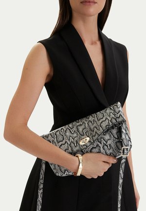 Donna con vestito nero senza maniche che tiene una pochette in pelle di serpente bianca e nera con chiusura dorata e un braccialetto coordinato al polso.