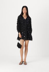 Vestido preto com aplicações de lantejoulas florais, design em V, mangas longas, finalizado com bordas frisadas. Combinado com uma bolsa preta e sandálias.