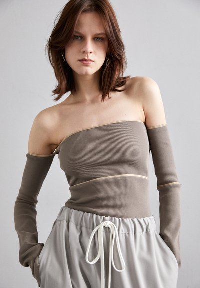 Ribbet, off-shoulder topp i taupe, med lange ermer og en kontrasterende gullsømdetalj. Kombinert med lys grå snortruser.