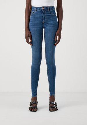 Modèle féminin portant un jean skinny bleu taille haute et des sandales noires à double bride avec boucle, debout contre un fond clair uni.