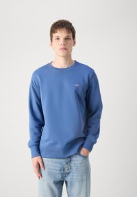 Young man wearing a blue sweatshirt and light blue jeans, standing with one hand in pocket against a plain background.