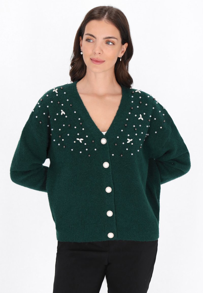 Zielony sweter z dzianiny z dekoltem w serek, ozdobiony dekoracyjnymi koralikami i zdobieniami. Posiada duże białe guziki z przodu.