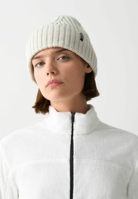 Krémová pletená čiapka so zloženým okrajom. Model má na sebe bielu fleece bundu s čiernym zipsom a vysokým golierom.