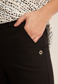 Czarne dopasowane spodnie wykonane z miękkiego materiału, z boczną kieszenią zdobioną złotym akcentem metalowym oraz gładkim, estetycznym designem.