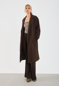 Una donna sta in piedi con le mani nelle tasche, indossando un lungo cappotto marrone scuro, un top senza spalline a stampa leopardata, pantaloni marrone scuro e scarpe a punta marroni.