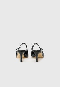 Schwarze Sandalen mit Absatz aus strukturiertem Lackleder, offener Rückseite und dünnem, gebogenem Absatz. Die Sohle hat eine helle, beige Farbe.