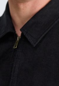 Zbliżenie na osobę w ciemnej kurtce z kordurowego materiału z widocznym częściowo zapiętym zamkiem błyskawicznym i kołnierzykiem.