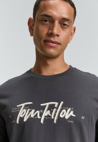 Camiseta de color gris oscuro hecha de algodón, con un cuello redondo, que presenta un logo y texto de color crema en el pecho en un estilo manuscrito.