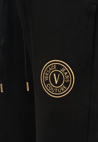 Μαύρο ύφασμα με χρυστό κεντημένο κυκλικό λογότυπο Versace Jeans Couture και μαύρα κορδόνια με χρυσά άκρα.