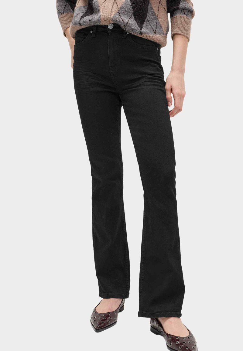 Personne portant un pantalon noir à jambe droite, un pull marron à motifs, et des ballerines pointues foncées avec des œillets, debout devant un fond clair.