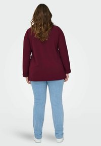 Burgundská halenka s dlouhým rukávem a ohrnutými manžetami, spojená s světle modrými džíny a bílými teniskami, zobrazená zezadu. Hladká textura látky.