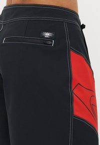 Crne kupaćice s crvenim detaljima, s pojasnim džepom na zatvarač, kontrastnim bijelim šavovima i logotipom. Glatka, lagana tkanina.