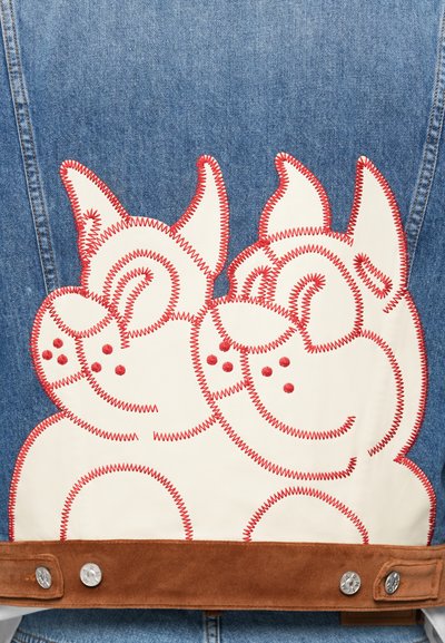 Τζάκετ από τζην με κόκκινες κεντητές απεικονίσεις δύο σκύλων σε κρεμ ύφασμα. Χαμηλότερο μέρος από καφέ σουέτ με μεταλλικά αξεσουάρ.