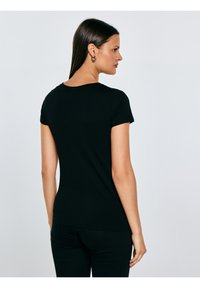 Hosszú sötét hajú nő arany karika fülbevalóval, testhezálló fekete rövid ujjú pólóban és fekete nadrágban, oldalra fordulva, egyszerű háttér előtt.