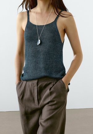 Femme portant un top sans manches en tricot gris foncé, un pantalon marron ample et un collier pendentif en argent, les mains dans les poches.
