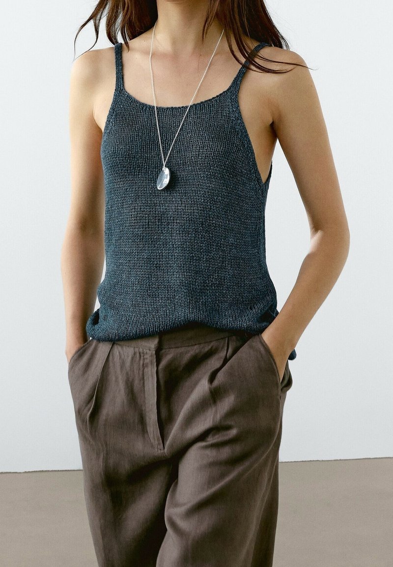 Femme portant un top sans manches en tricot gris foncé, un pantalon marron ample et un collier pendentif en argent, les mains dans les poches.