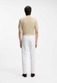 Hombre de pie de espaldas, vistiendo una camisa beige de manga corta, pantalones blancos y zapatos marrón oscuro sobre un fondo blanco.