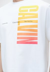 Λευκό μπλουζάκι με μεγάλο κατακόρυφο κείμενο με διαβάθμιση χρώματος "CALVIN" από κίτρινο σε ροζ και μικρότερο κείμενο "Calvin Klein New York City" και "Established 1968".